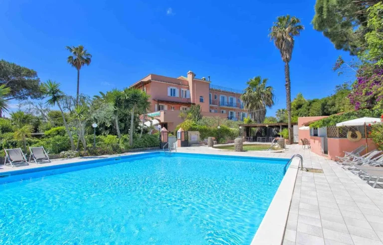 Piscina esterna semi-olimpionica con lettini, palme e la facciata rosa dell'hotel